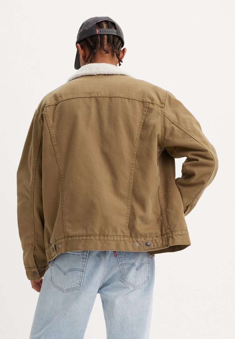 Veste en coton marron avec un col en sherpa blanc doux, coupe décontractée, coutures visibles et accents à boutons-pression, associée à un jean bleu clair.