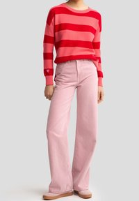 Červený a růžový pruhovaný svetr s žebrovanými manžetami, doplněný růžovými kalhotami se širokými nohavicemi a světle zbarvenými botami. Detaily srdce na manžetě svetru.