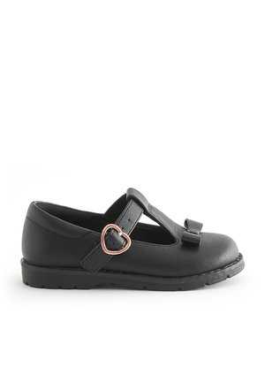Schwarzer Kinderschuh mit flacher Sohle und T-Riemen, herzförmiger goldener Schnalle und kleinem Schleifchen am Zehenbereich, dargestellt auf weißem Hintergrund.