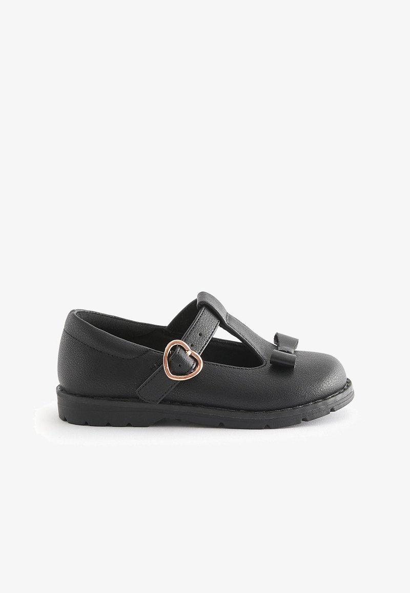 Chaussure plate noire pour enfants avec bride en T, boucle dorée en forme de cœur et petit nœud sur le bout, présentée sur fond blanc.