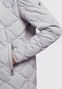 Veste matelassée gris clair avec une texture lisse, comportant une poche et un logo sur la manche. Des motifs arrondis créent un intérêt visuel.