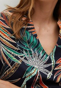 Halenka s krátkými rukávy a výstřihem do V, s tmavým pozadím a živými zelenými a oranžovými listovými vzory obrysovanými bílou, z texturované látky.