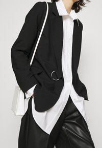 Černý blazer s detailem opasku, vrstvený přes bílou košili s knoflíky. V kombinaci s černými koženými kalhotami a bílou texturovanou kabelkou.