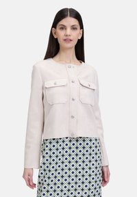 Hellbeige cropped Jacke mit zwei Fronttaschen, silbernen Knöpfen und einer glatten Textur. Kombiniert mit einem gemusterten Rock mit grünen und blauen Rauten.