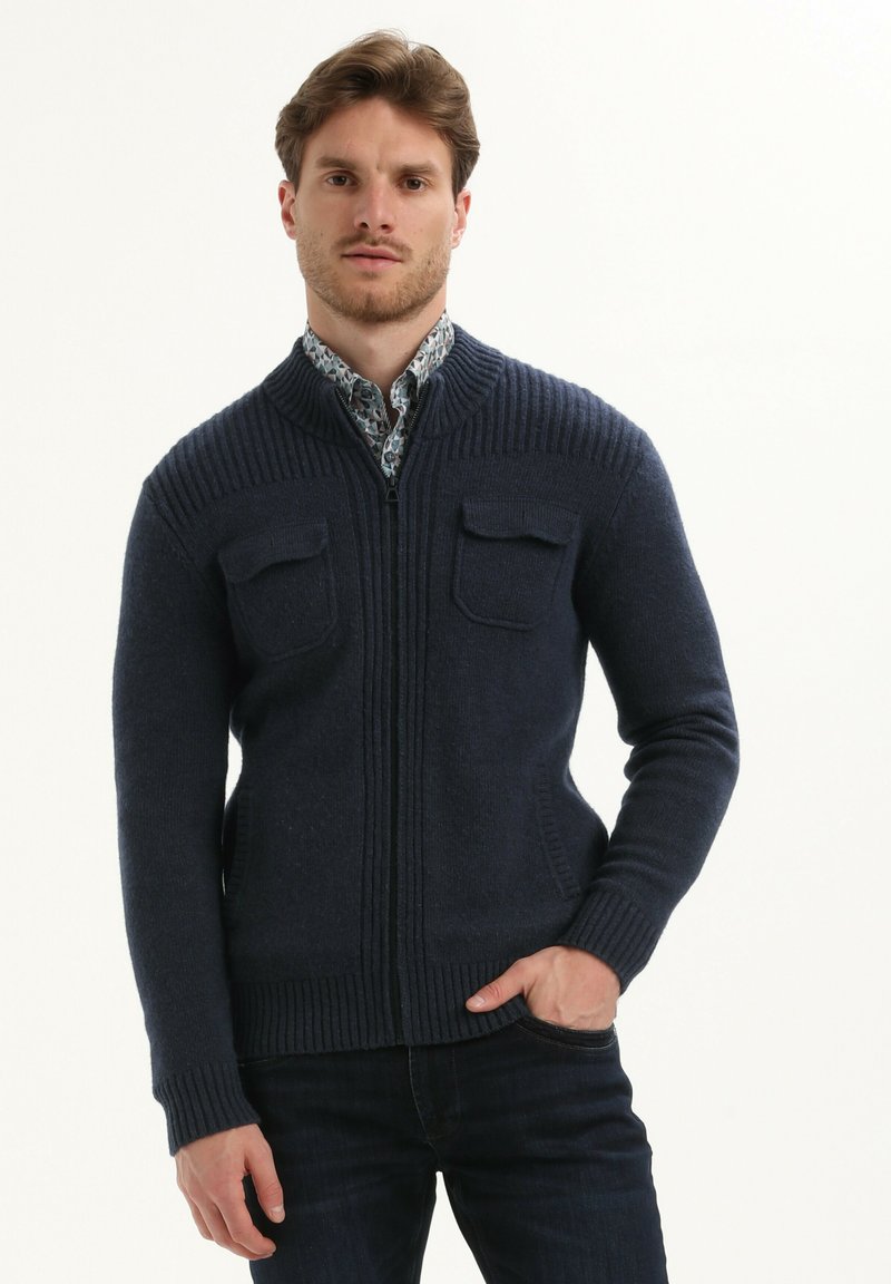 Cardigan a maglia blu navy con zipper intera, texture a coste, due tasche sul petto e colletto, indossato sopra una camicia con motivo.