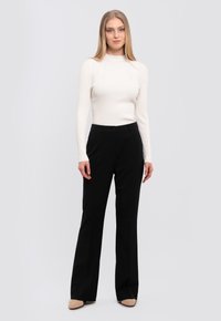 White ribbed long-sleeve top paired with black trousers, featuring a fitted design and flared leg. Shoes are beige pointy-toed. Plain background.