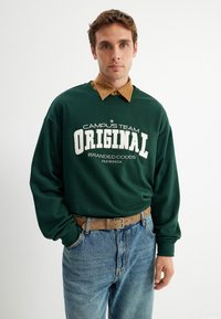Groene sweatshirt met de opdruk "CAMPUS TEAM ORIGINAL", gelaagd over een bruine overhemd met kraag, gecombineerd met blauwe jeans en een bruine riem.