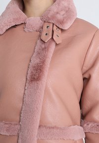 Růžová bunda z umělé kůže se světle růžovým límcem z umělé kožešiny. Má zatrhávací knoflíky a doplněk z umělé kožešiny vpředu.