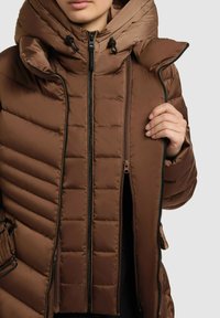 Chaqueta acolchada marrón con textura acolchada, cierre frontal con cremallera y una gran capucha desmontable. Presenta toggles y un bolsillo con cremallera.