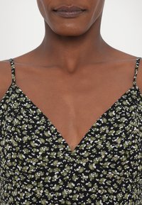 Robe noire à fleurs vertes et blanches, col en V et fines bretelles. Présente de petites décorations rondes sur le tissu.