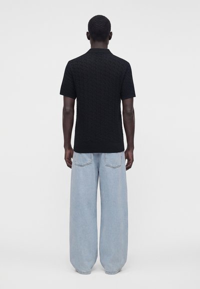 Homme vu de dos portant une chemise noire texturée à manches courtes et un jean large bleu clair ample, debout devant un fond uni.