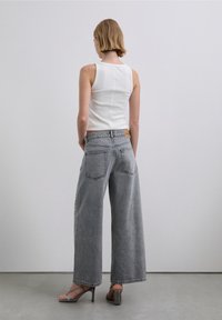 Bílý žebrovaný tílko spárované s kalhotami s vysokým pasem, širokými nohavicemi a šedou barvou. Kalhoty mají dvojité zadní kapsy a štítek u pasu.