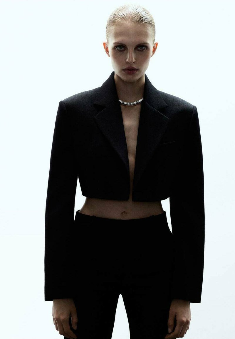 Blazer noir court avec épaules structurées, associé à un pantalon noir taille haute. Le mannequin porte un collier en chaîne argentée. Fond minimaliste.