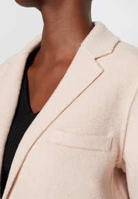 Gros plan d'une personne portant un manteau beige clair texturé sur un haut noir côtelé à col en V, montrant le cou et la partie supérieure de la poitrine.