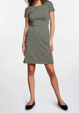 Femme portant une robe courte à manches courtes, longueur genou, avec un motif géométrique répétitif et des chaussures plates noires, debout devant un fond uni.