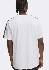 Uomo con capelli intrecciati che indossa una maglietta bianca a maniche corte con nastro nero con logo lungo le spalle e le maniche, visto di spalle.