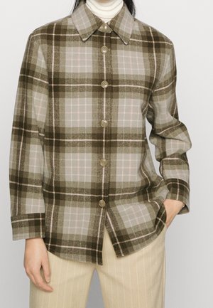 Woman wearing brown and beige plaid button-up shirt over white turtleneck, paired with light beige pinstriped pants, hand in pocket.