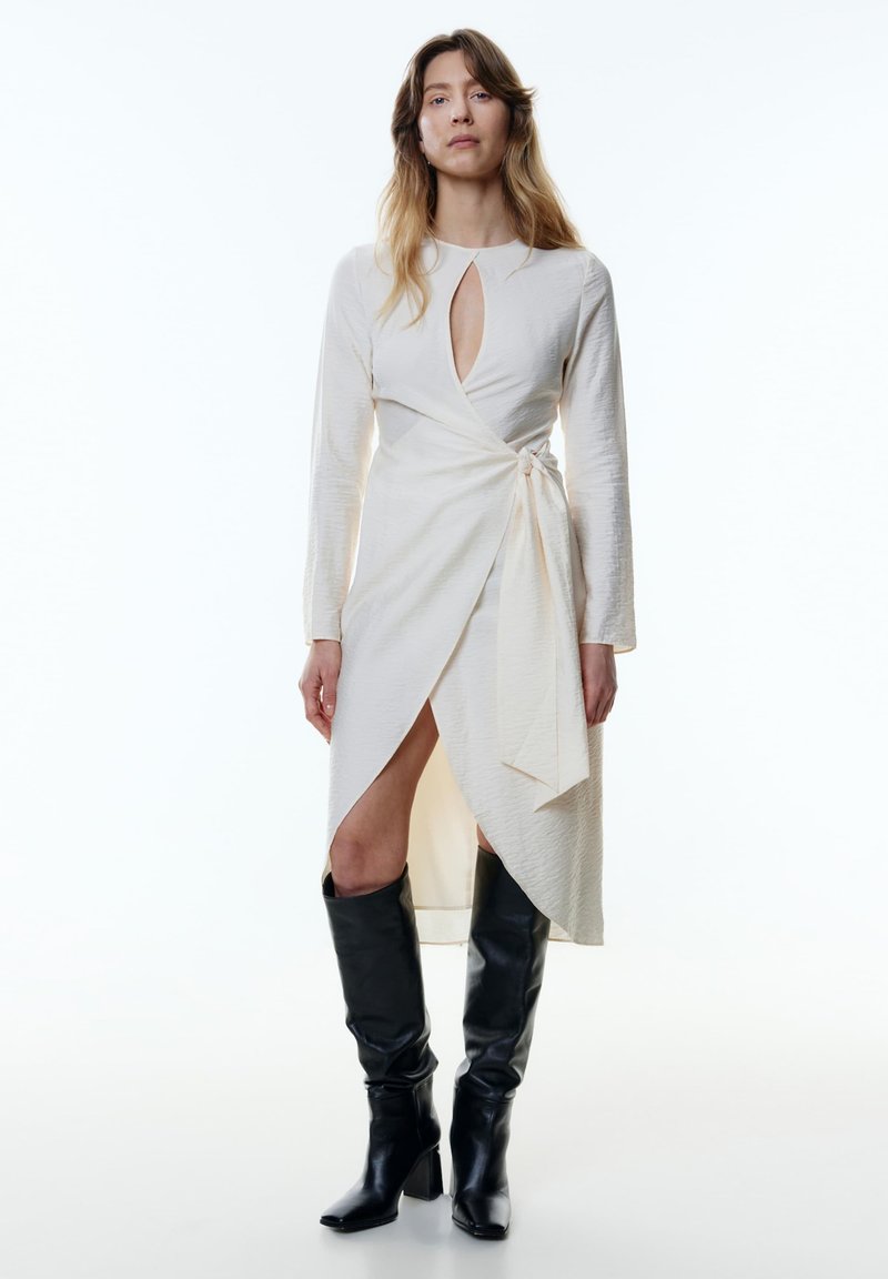 Langärmliges, cremefarbenes Wickelkleid mit einem Schlüssel-Ausschnitt und seitlicher Bindung; kombiniert mit knielangen schwarzen Lederstiefeln im quadratischen Zehendesign.