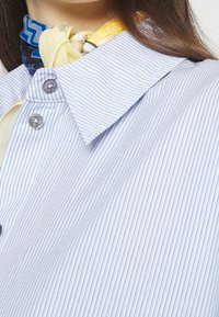 Camicia a righe blu con colletto a punta e due bottoni, realizzata in un tessuto testurizzato. Una sciarpa a fantasia gialla e blu è annodata intorno al collo.