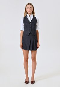 Gilet in denim scuro con un design aderente e chiusura a bottoni; abbinato a una camicia bianca con bottoni e una gonna plissettata. Tacchi a punta marroni.