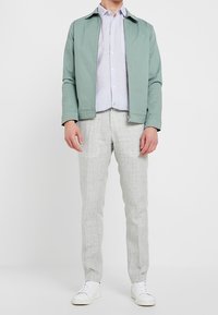 Giacca zip-up verde chiaro sopra una camicia azzurra chiara, abbinata a pantaloni grigi chiari e sneakers bianche. Texture liscia, design casual.