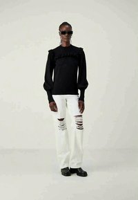 Top preto de manga longa com decote franzido, combinado com jeans brancos de perna ampla e rasgada. O modelo usa óculos de sol escuros e botas pretas robustas.