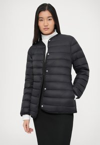 Kvinne iført en svart quiltet jakke over en hvit turtleneck og svart skjørt, stående mot en ensfarget lys bakgrunn.