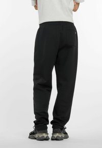 Pantalons de survêtement noirs avec une taille élastique, présentant une poche arrière et des chevilles resserrées, associés à des baskets noires et grises sur un fond uni.