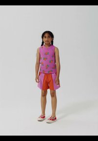 Mädchen mit dunklem geflochtenem Haar trägt ein ärmelloses lila Oberteil mit Fruchtmustern, zweifarbige Shorts in Pink und Orange sowie rote Turnschuhe.