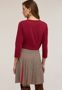 Vínová dlouhá halenka s kulatým výstřihem, která je kombinována s plisovanou sukní v houndstooth vzoru s červenými akcenty a siluetou fit-and-flare.