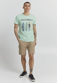 Miętowa bawełniana koszulka z grafiką deski surfingowej oraz napisem "CALIFORNIA", w zestawie z beżowymi szortami cargo i czarnymi sneakersami.