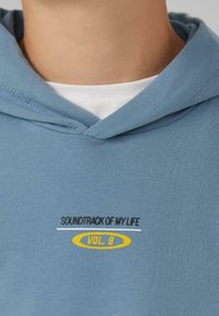 Helderblauwe katoenen hoodie met een buidelzak, trekkoordcapuchon en geborduurde tekst "SOUNDTRACK OF MY LIFE VOL. 8" in zwart en geel.