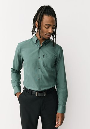 Mężczyzna z dredami w zielonej koszuli zapinanej na guziki i czarnych spodniach, stojący z jedną ręką w kieszeni, patrzący w dół.