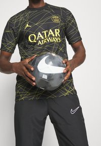 Maglia nera con linee geometriche gialle e scritta "Qatar Airways". Tiene in mano un pallone da calcio Nike grigio e nero, indossa pantaloni scuri.