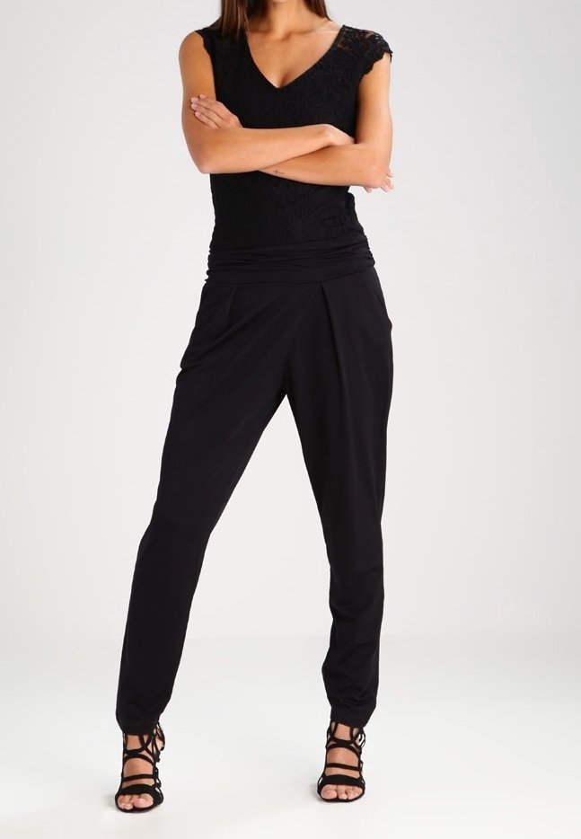 Femme debout, les bras croisés, portant un top sans manches en dentelle noire, un pantalon plissé taille haute noir et des talons aiguilles striés noirs.