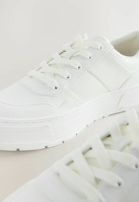 Sneakers bianchi con tomaia sintetica testurizzata, dotate di lacci piatti, punta rotonda e suola spessa in gomma. Accentuati da pannelli cuciti.