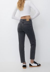 Jeans negros de tiro alto con cinco bolsillos y una etiqueta de cuero marrón, combinados con zapatillas negras y blancas, sobre un fondo liso.