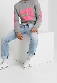 Sweatshirt gris avec logo "Levi's" rose et vert, associé à un jean bleu clair et des baskets blanches. Le sweatshirt a des poignets et un col côtelés.