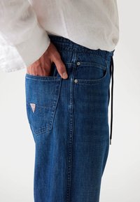 Blue denim pants with an elastic waist and drawstring, featuring a back pocket with a triangular logo patch. Fabric has a smooth texture.