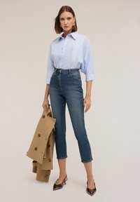 Camicia azzurra a righe con bottoni, jeans scuri a vita alta, e un trench coat beige, insieme a scarpe col tacco marroni lucide e accessori.