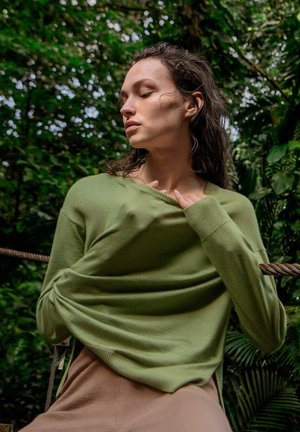 Kvinde med lukkede øjne trækker grøn sweater over beige bukser, omgivet af tæt grønt løv udendørs.