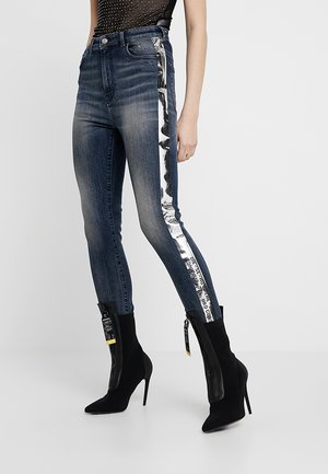 Slim-Fit ausgebleichte blaue Jeans mit reflektierenden silbernen Streifen und schwarzem Text an den Seiten, kombiniert mit schwarzen hochhackigen Stiefeletten mit Reißverschlüssen.