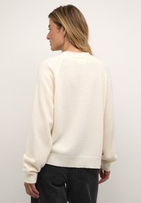 Woman facing sideways wearing a cream knit sweater with long sleeves and black pants against a plain background.
