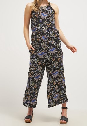 Frau trägt einen ärmellosen schwarzen Jumpsuit mit blauen und orangefarbenen Blumenmustern und schwarzen offenen Zehen Knöchelriemen-Sandalen, steht locker.