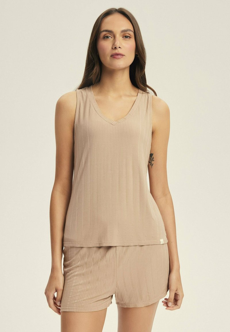 Woman wearing beige ribbed sleeveless top and matching shorts standing against plain background, tattoo visible on left arm.