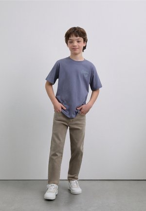 Camiseta morada de manga corta con logo impreso, combinada con jeans beige y zapatillas blancas sobre un suelo gris y fondo liso.