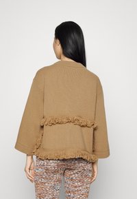 Cardigan marron tricoté avec manches courtes, présentant des détails texturés avec franges le long de l'ourlet. Porté avec des leggings à motifs.