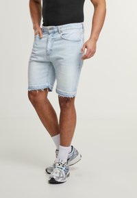 Világoskék farmer sort, kézzel szaggatott szegéllyel, öt zsebes dizájnnal és gombos zárással. Fehér zoknikkal és ezüst futócipőkkel párosítva.