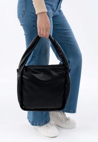 Borsa tote in pelle nera con una superficie liscia e una maniglia arrotondata, dotata di chiusura zip nella parte superiore. Indossata con jeans in denim e sneakers bianche.
