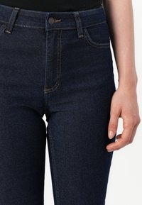 Σκούρο μπλε τζιν παντελόνι φορεμένο με μαύρα ρούχα, που δείχνει μπροστινές τσέπες, λεπτομέρειες ραφής και ένα χέρι να ακουμπάει στο μηρό.